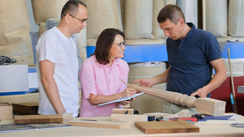 A design professional holding a piece of wood furniture talks with two individuals in a facility.