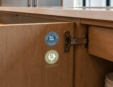 Open wood kitchen island cabinet door showing KCMA “Quality Tested” and “Responsible Sustainable” certification stickers, with stacked bowls inside.