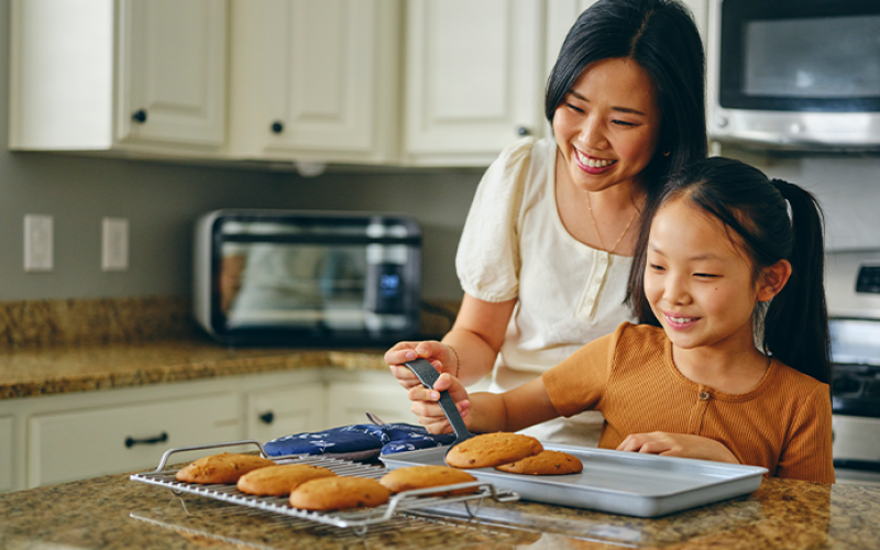 A mother and daughter baking in their kitchen with white cabinets in the background.