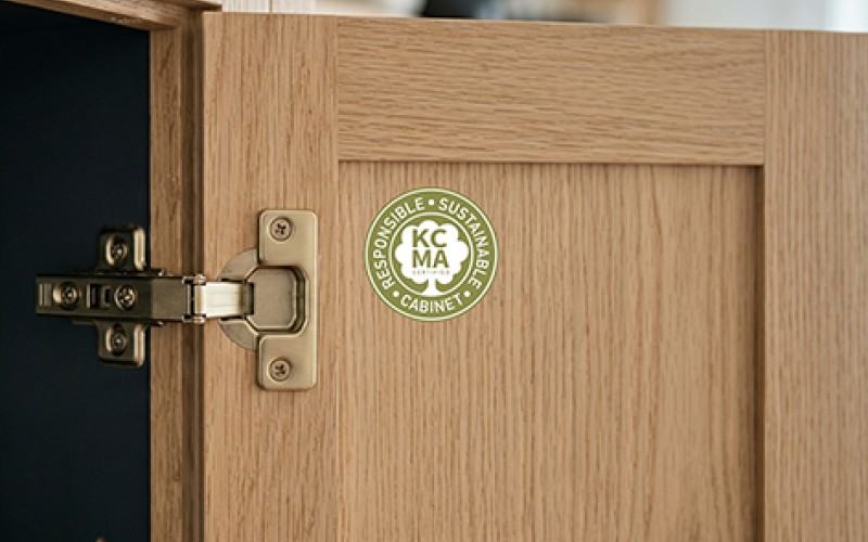 Close-up of a light wood kitchen cabinet door featuring a green KCMA "Responsible Sustainable Cabinet" certification seal on the interior panel.