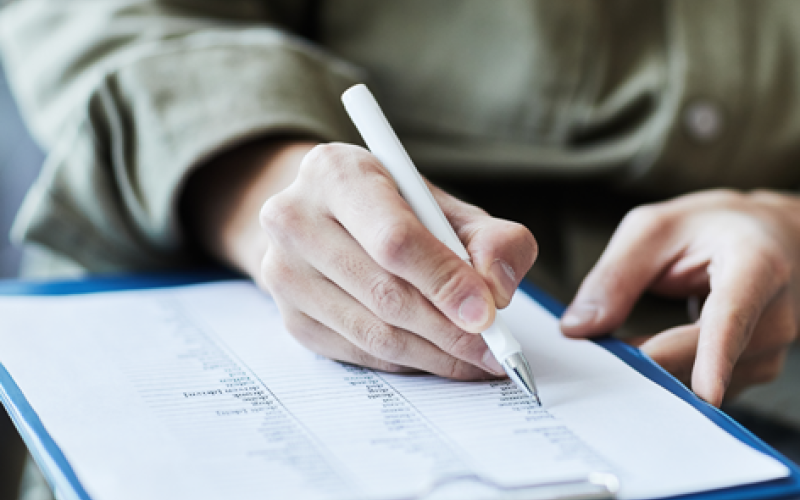 A close-up of a person's hand using a white pen to fill out a document on a blue clipboard, symbolizing the KCMA ESP application process.